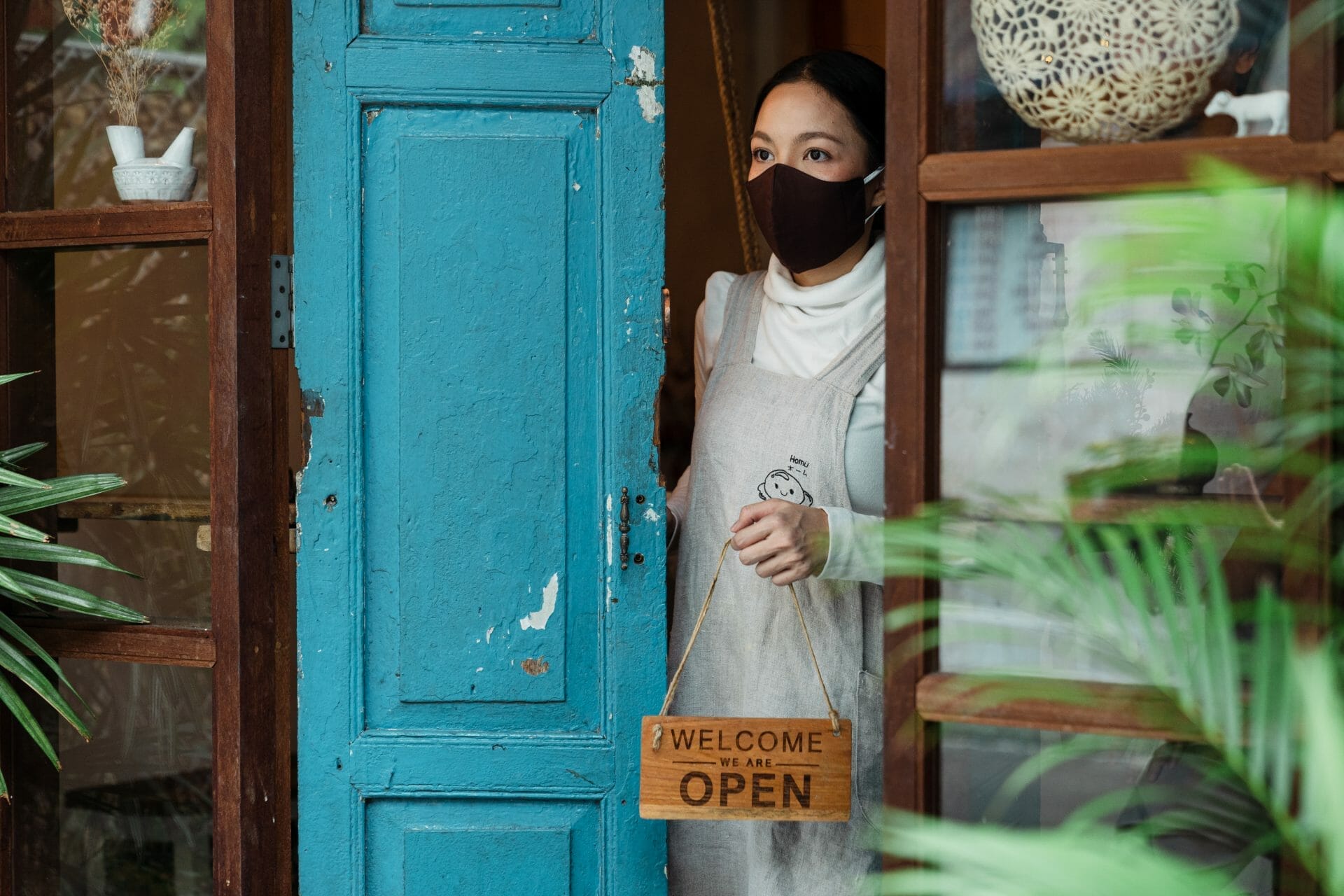 A woman wearing a mask and holding a sign opens her shop